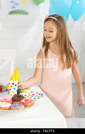 Adorable kid al tavolo di compleanno con decorazioni di festa e ciambelle durante il party Foto Stock