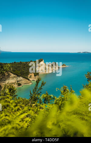 Cape Drastis (Perulades) con acque blu cristallo del mare su una soleggiata giornata estiva con foglie in primo piano (Corfu, Grecia, Europa) Foto Stock