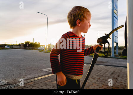Bambino tenere il tubo di una pompa del carburante al tramonto in corrispondenza di una stazione di gas. Foto Stock