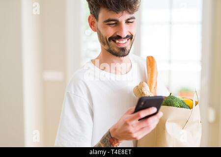 Giovane uomo bello tenendo un sacchetto di carta pieno di negozi di generi alimentari freschi a casa, mostrando e tramite smartphone durante l acquisto di prodotti Foto Stock