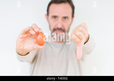 Senior uomo con uovo fresco su sfondo isolato con volto arrabbiato, segno negativo che mostra antipatia con il pollice verso il basso, il concetto di rifiuto Foto Stock