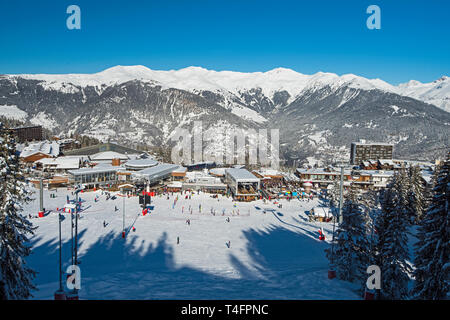 Vista panoramica verso il basso coperto di neve valle nella catena montuosa alpina con ski resort village center sul cielo blu sullo sfondo Foto Stock