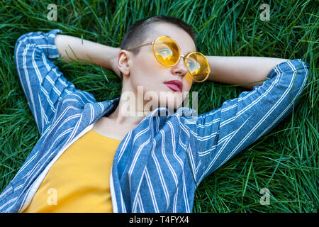 Ritratto di pensosa giovane donna con capelli corti in casual blue tuta a strisce, maglietta gialla, bicchieri sdraiati sull'erba verde, tenendo le mani dietro Foto Stock