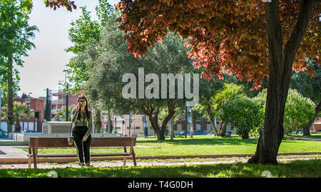 Felice lonely donna o ragazza seduto su una panchina nel parco sorridente in autunno o caduta stagione in attesa di qualcuno e utilizzando il suo telefono, Murcia, Spagna, 2019. Foto Stock