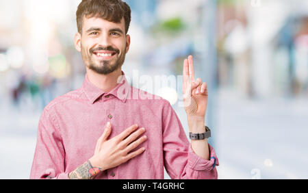 Giovane uomo bello indossare maglia rosa su sfondo isolato il giuramento con la mano sul petto e sulle dita, facendo una promessa di fedeltà giurata Foto Stock