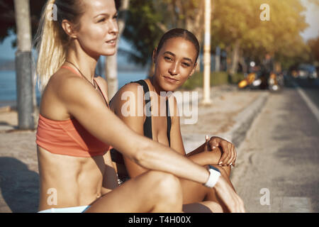 Montare la giovane donna in sportswear sorridere mentre seduto su un marciapiede con il suo partner di allenamento prendendo una pausa dalla loro esecuzione Foto Stock