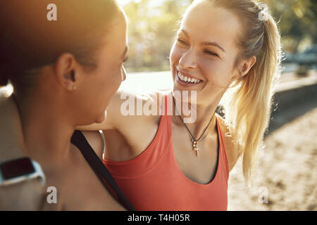 Due ridere delle giovani donne in abbigliamento sportivo in piedi a braccetto assieme al di fuori in una giornata di sole prima di correre insieme Foto Stock