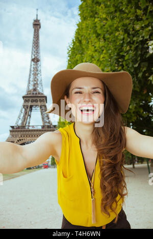 Sorridente giovane turista donna in giallo camicetta e hat tenendo selfie contro chiara vista della Torre Eiffel a Parigi, Francia. Foto Stock