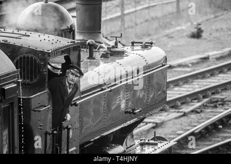 Black & White primo piano della locomotiva a vapore britannica d'epoca sul binario ferroviario in arrivo alla stazione; autista dell'equipaggio del treno a vapore in cabina in cabina in cabina, guardando in alto. Foto Stock