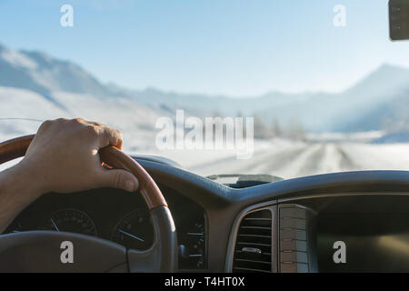 Close-up di una mano di un uomo al volante di una vettura che si muove su una strada innevata tra le montagne in inverno Foto Stock