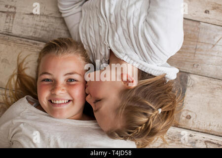 Ritratto di una ragazza baciato sulla guancia con la sua sorella più giovane mentre giaceva su un pavimento di legno. Foto Stock