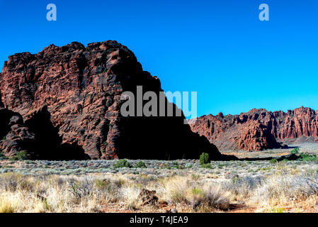 Vasto deserto campi di sagebrush e cespugli verdi con alte e ripide rossastro formazioni rocciose e montagne sotto un cielo blu. Foto Stock