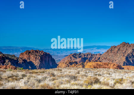 Backcountry deserto di campi con cespugli di morti e spazzola, ripide e frastagliate colline rocciose e montagne sotto un cielo blu. Foto Stock