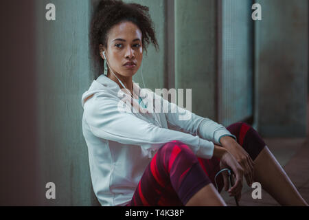 Donna seduta da strada appoggiata ad un pilastro e guardando lontano. Urban runner riposo dopo la sessione di allenamento. Foto Stock