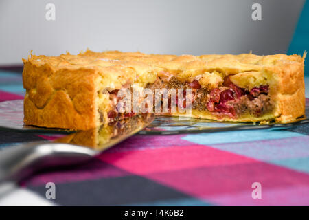 Sostenuto croccanti e dolci fatti in casa torta di ciliegie su una lastra di vetro e una tabella coperta con colorati cubo panno pattern close up Foto Stock