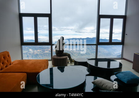 Camera in hotel con paesaggio di montagna vista da finestra, interni con tavolo e sedie e un uomo si siede a vedere la scena attraverso la finestra Foto Stock