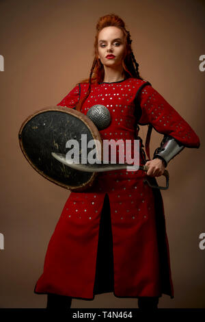 Brave, gravi donna che indossa come nobile eroe in rosso tunica medievale. Bello, affascinante modello con lo zenzero capelli tenendo scudo e pugnale, ponendo in studio con arma. Foto Stock