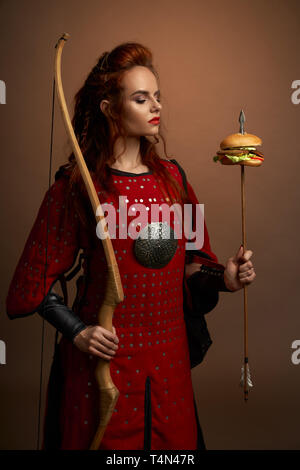 Medievale guerriero femmina tenendo la prua e la freccia con hamburger, guardando a gustosi piatti fast food Burger. Donna Gorgeous in tunica rossa con arma in posa e in piedi in studio. Foto Stock