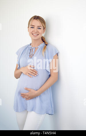 Gravidanza felice giovane donna cullano il suo ventre con le sue mani come ha legami con il suo bambino in posa contro un muro bianco sorridente al camer Foto Stock