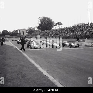 Anni sessanta, storico motor racing al Crystal Palace il circuito di gara nel sud di Londra, London, England, Regno Unito, piloti e vetture in pista, in posizione sulla griglia di avvio, con un ufficiale di gara tenendo in mano un cartello che diceva 1 min fino alla partenza della gara. Foto Stock