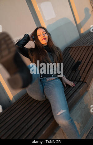 Giovane donna ascolta la musica nelle cuffie chiuse attraverso il suo telefono che indossa una giacca di pelle e jeans in corrispondenza di un tramonto vicino al fiume Daugava Foto Stock
