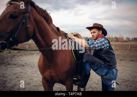 Brutale cowboy in jeans e giacca di pelle si arrampica a cavallo su ranch del Texas occidentale. Vintage persona di sesso maschile con cavallo selvaggio west Foto Stock