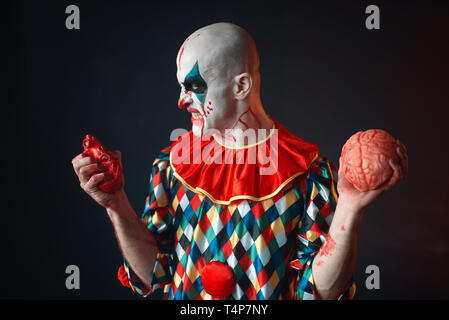 Mad sanguinosa clown detiene il cuore umano e il cervello. L'uomo con il trucco in costume di carnevale, crazy maniac Foto Stock