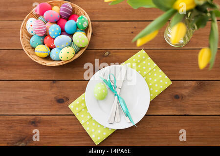 Pasqua, vacanze e concetto di oggetto - uova colorate nel cestello, piatti, posate e fiori sul tavolo di legno Foto Stock