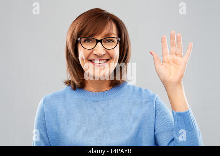 Gesto, visione e il vecchio concetto di persone - Ritratto di sorridere senior donna in bicchieri agitando la mano su uno sfondo grigio Foto Stock