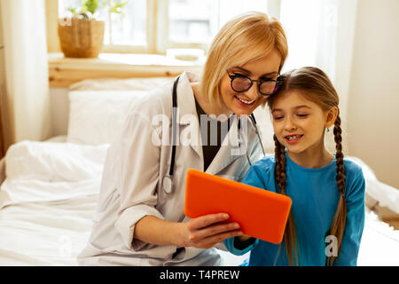 Trascorrere il tempo con un medico. Trasmissione via IR di allegro attraente bella medico che indossano occhiali guardando la compressa arancione lungo con una bella carino Foto Stock