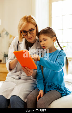 La lettura di informazioni in un tablet. Dolce abbastanza interessato i capelli castani ragazza puntando al tablet mentre un affascinante attraente bionda grave medico donna in Foto Stock