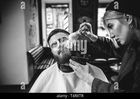 Client durante la rasatura della barba in barberia. Barbiere femmina al salone. La parità tra i sessi. Donna nel maschio professione. Nero e bianco o incolore foto. Acconciatura, salone di bellezza e parrucchiere, il concetto di stile di vita. Foto Stock