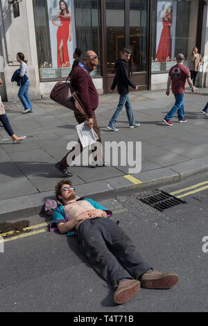 I passanti da guardare in giù su un giovane attivista in appoggio con la sua testa su un Oxford Circus frenare il giorno 4 di proteste da parte del cambiamento climatico attivisti ambientali con il gruppo di pressione la ribellione di estinzione, sul diciottesimo mese di aprile 2019, a Londra, in Inghilterra. Foto Stock