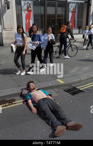 I passanti da guardare in giù su un giovane attivista in appoggio con la sua testa su un Oxford Circus frenare il giorno 4 di proteste da parte del cambiamento climatico attivisti ambientali con il gruppo di pressione la ribellione di estinzione, sul diciottesimo mese di aprile 2019, a Londra, in Inghilterra. Foto Stock