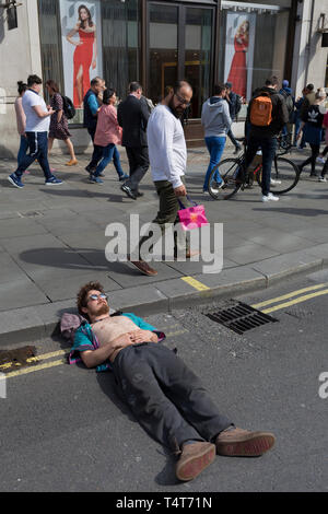 I passanti da guardare in giù su un giovane attivista in appoggio con la sua testa su un Oxford Circus frenare il giorno 4 di proteste da parte del cambiamento climatico attivisti ambientali con il gruppo di pressione la ribellione di estinzione, sul diciottesimo mese di aprile 2019, a Londra, in Inghilterra. Foto Stock