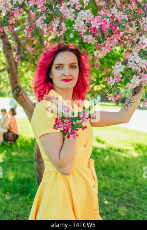 Bella e giovane donna con luminosi capelli rossi e ref labbra in piedi accanto a una fioritura di melo in un abito giallo. Foto Stock