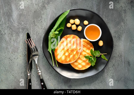 Grigliata di fettine rotonde di formaggio greco con miele, fumo-formaggio essiccato, aglio selvatico, menta e foglie di coriandolo. Lay piatto sul tavolo a trama. Foto Stock