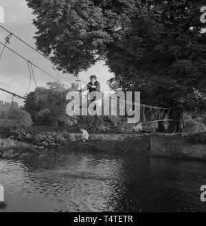 Anni sessanta, storico, pioggia si riversa giù come un giovane scout avventura attraversa un fiume su un ponte di corde gli scout hanno recentemente realizzato e legato ad un albero sul greto del fiume, Inghilterra, Regno Unito. Foto Stock