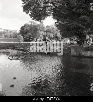 Anni sessanta, storico, un giovane scout di avventura e team attraversare un fiume su un ponte di corde gli scout hanno recentemente realizzato e legato ad un albero sul greto del fiume, Inghilterra, Regno Unito. Foto Stock