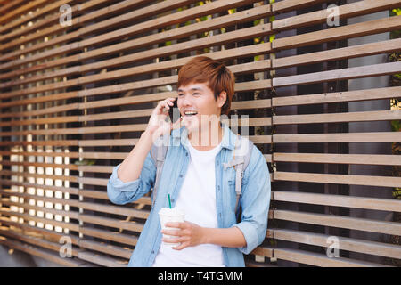 Giovane uomo asiatico sorridente e parlando al telefono mobile esterno con spazio copia sulla sinistra Foto Stock