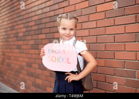 Piccola ragazza bionda in uniforme scolastica sorge vicino a un muro di mattoni in possesso di una lavagna con il testo torna a scuola. Foto Stock