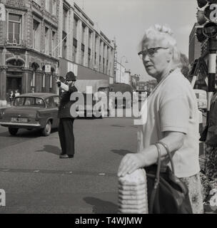 Anni sessanta, storici British poliziotto in uniforme e casco in piedi dirigendo il trafffic in un affollato South London street, con un pedone aldy al semaforo in trepidante attesa per attraversare la strada. Foto Stock