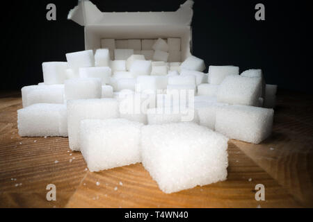 Macro shot di cubetti di zucchero accanto alla scatola di cartone sul tavolo di legno. Cibo malsano dolcificante, dolci cubetti di cristallo Foto Stock