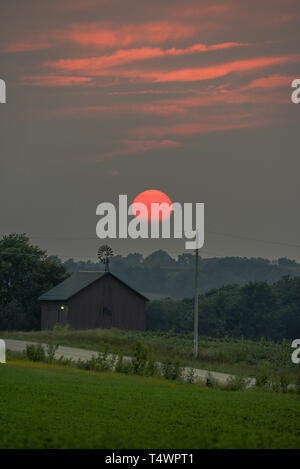 Afoso tramonto sul fienile rurale e mulino a vento lungo la strada di campagna in estate, Monroe, Wisconsin, STATI UNITI D'AMERICA Foto Stock