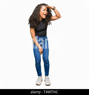 Giovane bella donna con capelli ricci molto felice e sorridente guardando lontano con la mano sopra la testa. Concetto di ricerca. Foto Stock