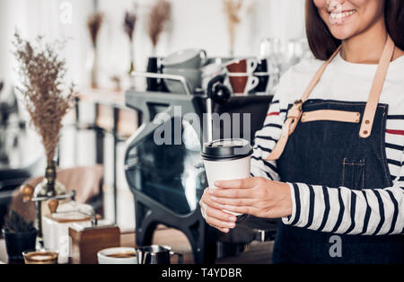 Barista serve prendere awary tazza di caffè presso il cafe.femmina asiatica barista indossare jean grembiule azienda caffè servito al cliente con volto sorridente al banco bar, Foto Stock