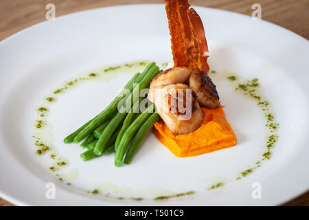 Cucinato al momento capesante su un letto di patate dolci serviti con fagiolini, bacon croccante e salsa verde su una piastra bianca in un ristorante Foto Stock