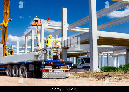 Lavoratore sta preparando il gancio della gru per lo scarico di un travetto in cemento da camion rimorchio. Foto Stock