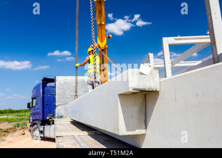 Lavoratore sta preparando il gancio della gru per lo scarico di un travetto in cemento da camion rimorchio. Foto Stock