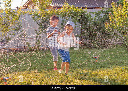 Attività estive. I bambini a giocare all'aperto con impianto automatico sistema di irrigazione. Ragazzo sorridente avendo divertimento all'aperto. I tubi flessibili di acqua di abbeveraggio l'erba Foto Stock